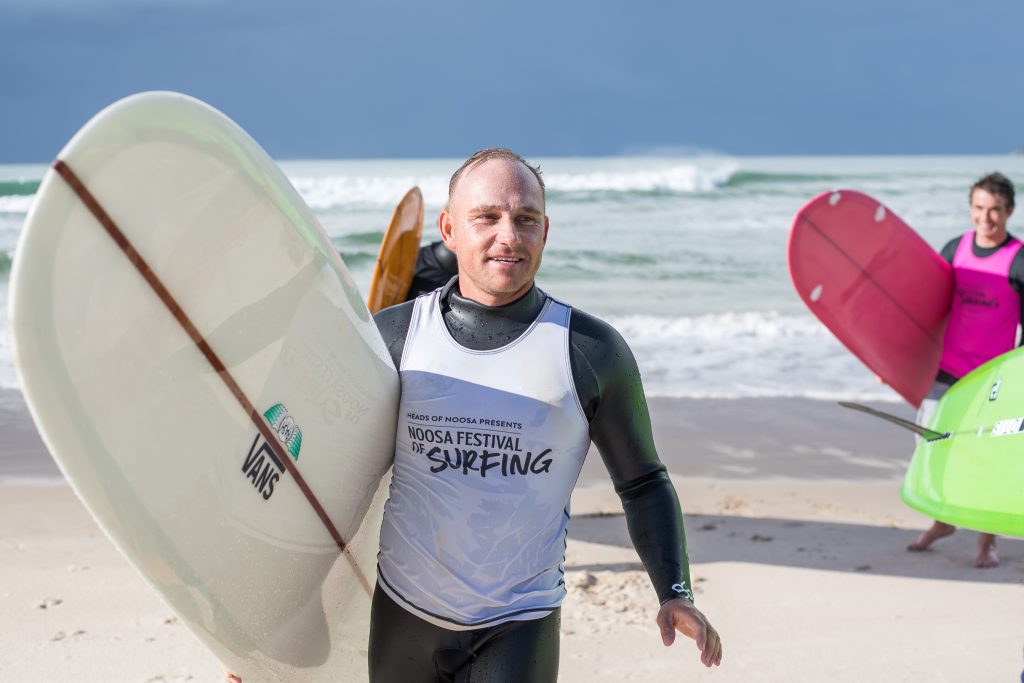 A man in a wetsuit and "Noosa Festival of Surfing" vest carries a surfboard on a sandy beach, with waves in the background and other surfers holding surfboards nearby.
