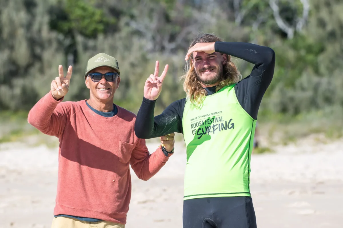 Two men stand on a sandy beach, both smiling and flashing peace signs. One wears a green surfing shirt and wetsuit, the other wears sunglasses, a cap, and a rust-colored sweater. Trees are blurred in the background.