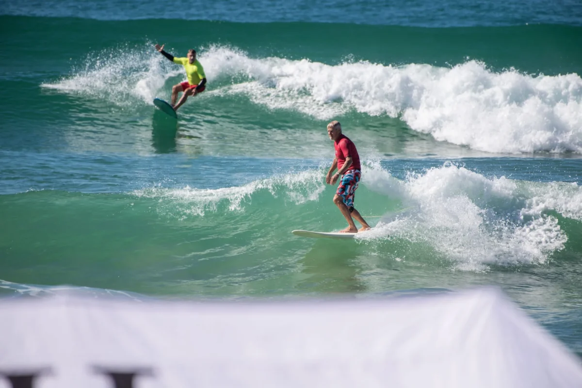 Two surfers ride ocean waves, one in a red shirt and patterned shorts in the foreground, and another in a yellow shirt further back. A blurred white object is visible at the bottom of the image.