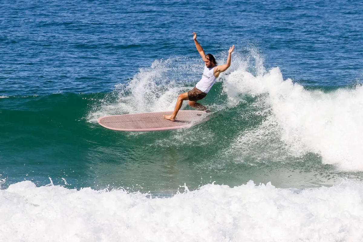 A person in a white tank top surfs a wave on a longboard, arms raised for balance, with blue ocean water and white surf in the background.