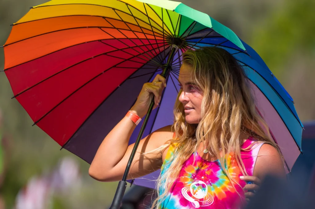 A woman with long blonde hair stands outdoors, holding a large rainbow umbrella. She wears a colorful tie-dye shirt and looks to the side, bathed in sunlight.