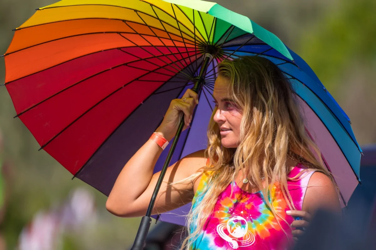 A woman with long blonde hair stands outdoors, holding a large rainbow umbrella. She wears a colorful tie-dye shirt and looks to the side, bathed in sunlight.