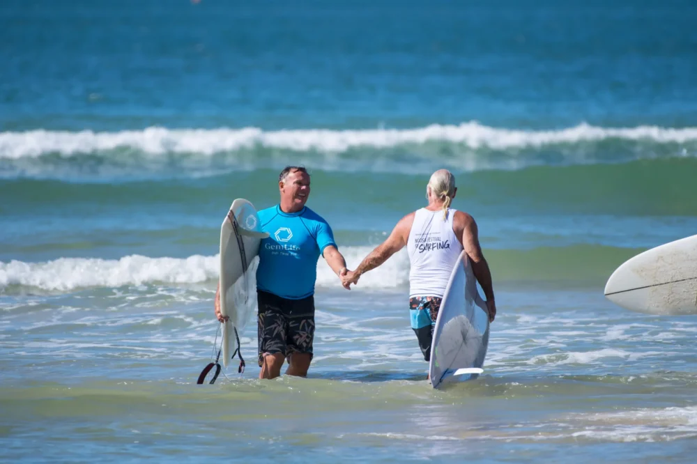 Two older men holding hands and smiling as they walk through shallow ocean water with surfboards, wearing casual surf attire, with waves and blue sky in the background.