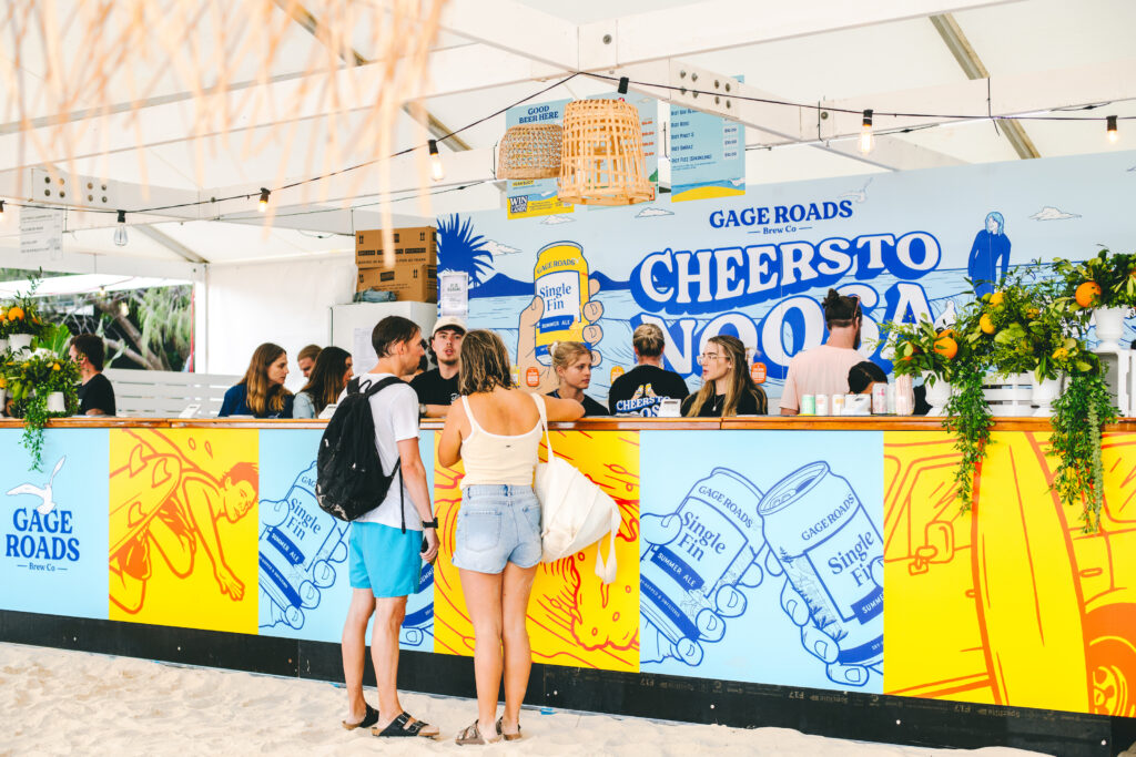 People order drinks at a colorful beachside bar decorated with vibrant artwork, plants, and signs reading "Cheers to No Drama" and "Gage Roads." The area is bright, lively, and set on sand.