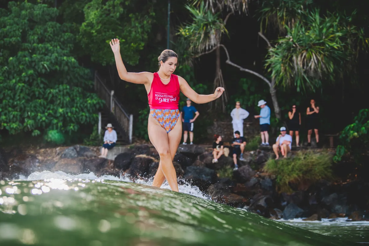 A woman in a red tank top and colorful shorts balances on a surfboard, riding a wave near rocks and lush greenery, while several people watch from the shore in the background.