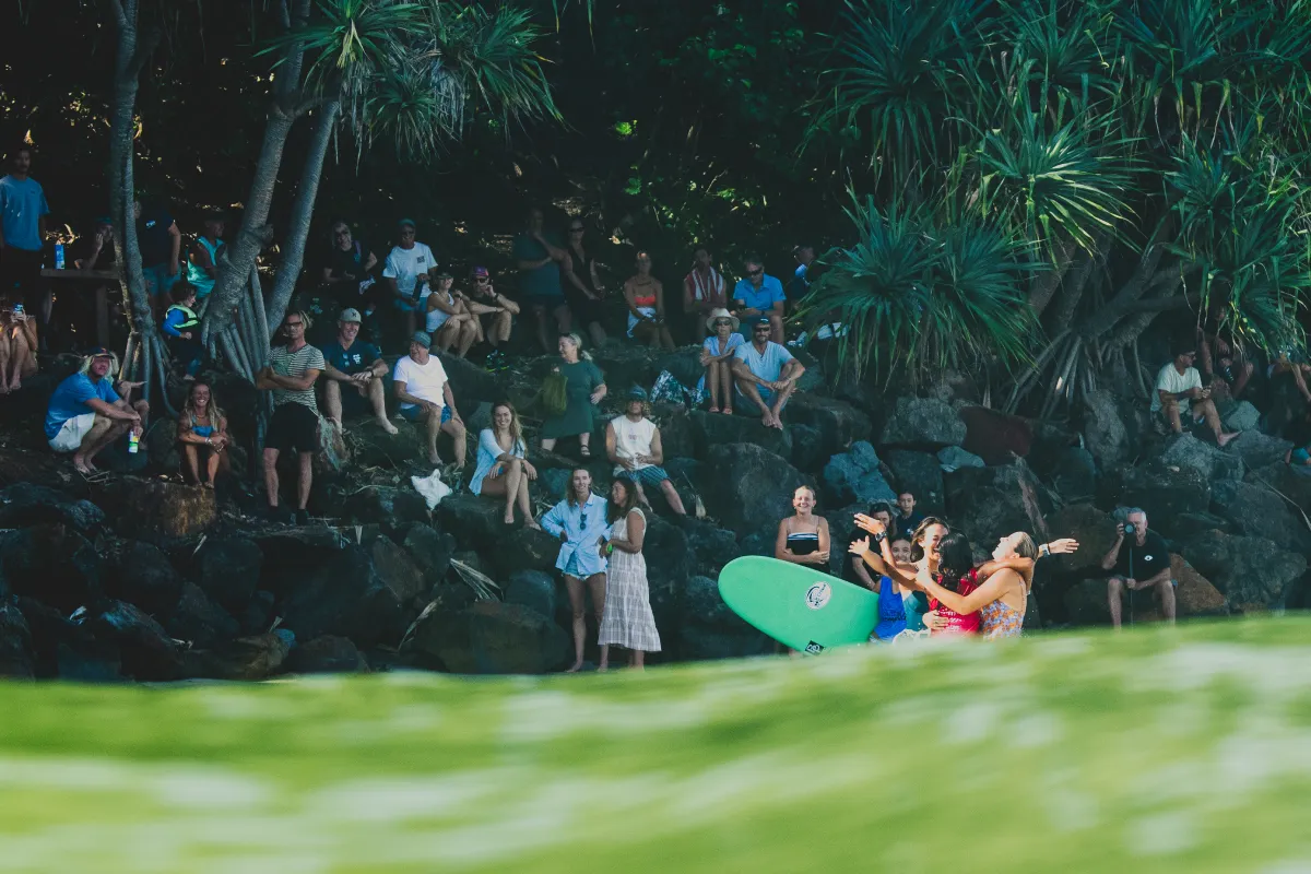 A group of people with surfboards celebrate near the shore, while a crowd sits on rocks and grass under trees in the background, watching the scene.