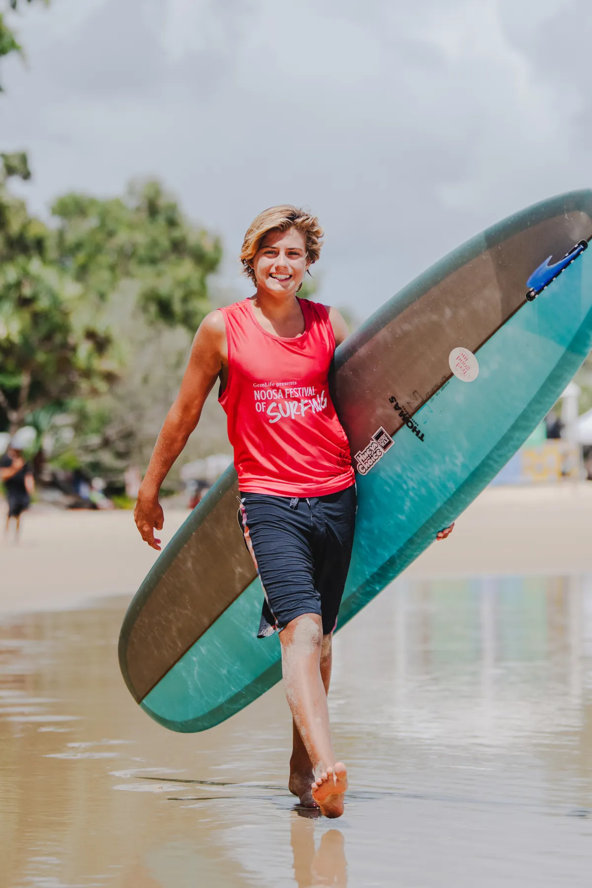 A smiling young person in a red tank top and black shorts walks barefoot on a wet beach, carrying a blue surfboard under one arm. Trees and a cloudy sky are visible in the background.