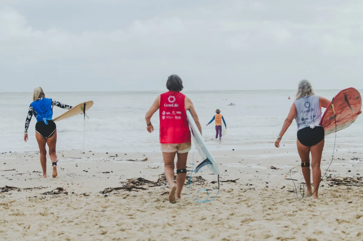 Three people holding surfboards walk toward the ocean from a sandy beach, with another person in the water ahead of them. The sky is overcast and the beach is scattered with seaweed.