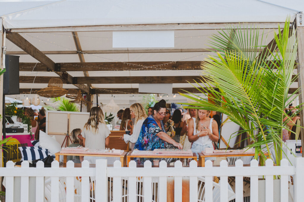 A lively outdoor event under a large tent, with people socializing around tables and tropical plants, festive decorations, and a white picket fence in the foreground.
