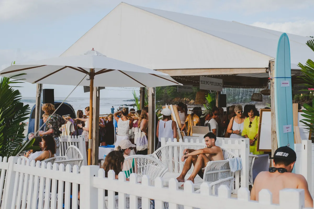 A crowd gathers under a white tent at a beach market. People browse stalls while others relax on white chairs near a white picket fence. Surfboards and palm leaves add to the tropical atmosphere by the sea.