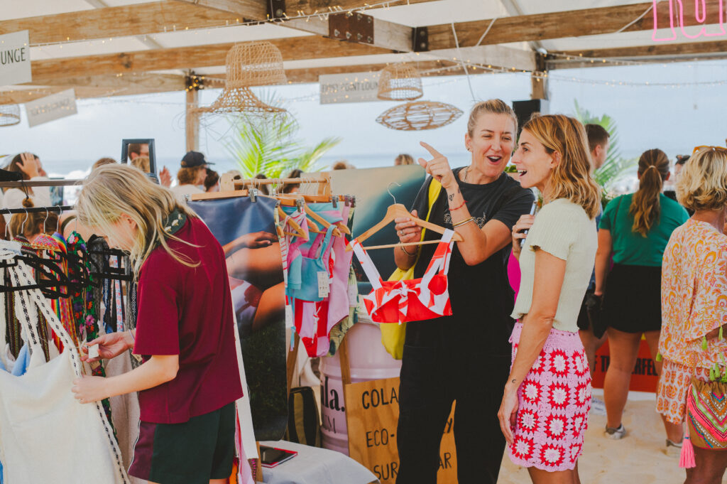Two women are smiling and talking while holding swimsuits at a beachside market stall, surrounded by racks of colorful swimwear. Other shoppers browse in the background under a wooden canopy decorated with lights.