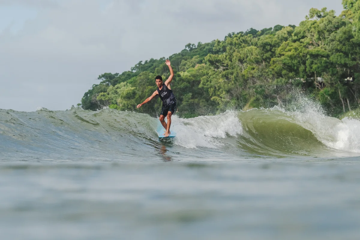 A man in a black wetsuit surfs on a small wave near a forested shoreline, balancing with one arm raised under a cloudy sky.