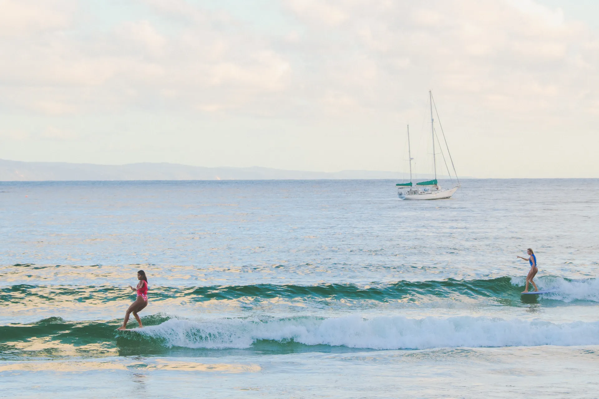 Two people surf small waves near the shoreline, while a white sailboat floats in the calm ocean in the background under a partly cloudy sky.