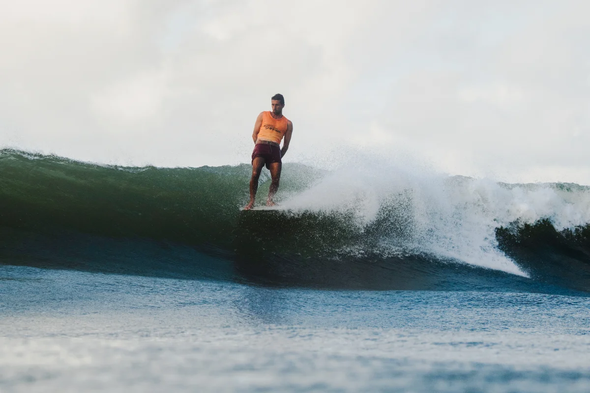 A surfer wearing a yellow-orange shirt and red shorts rides on the nose of a dark green wave, balancing with arms by his sides under a cloudy sky.