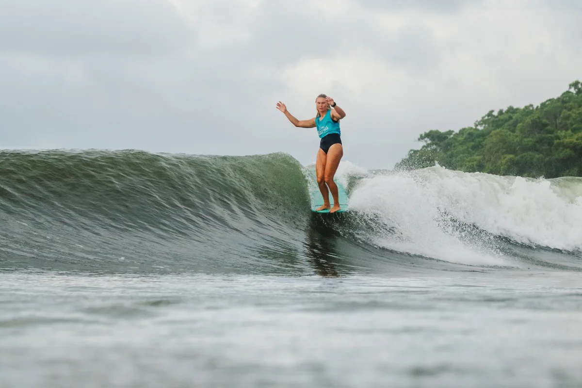 A person in a blue swimsuit rides a surfboard on the crest of a wave, arms outstretched for balance. Lush green trees and a cloudy sky are visible in the background.