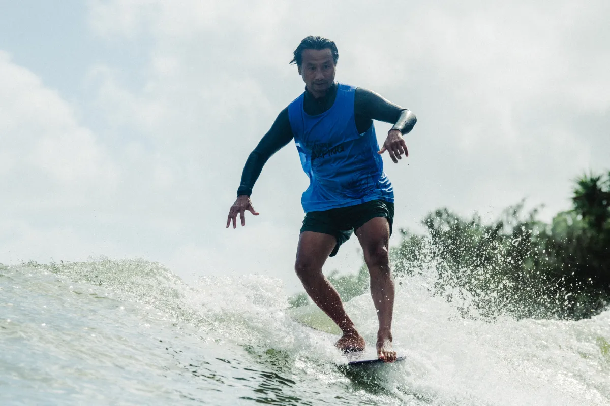 A man wearing a blue tank top surfs on a wave, balancing with bent knees and outstretched arms, with water splashing around his feet and trees visible in the background.