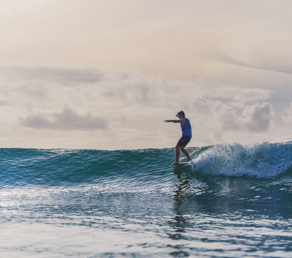 A person in blue shorts and a tank top surfs on a small wave, balancing on a surfboard with arms outstretched. The ocean is calm, and the sky is partly cloudy with soft, warm light.