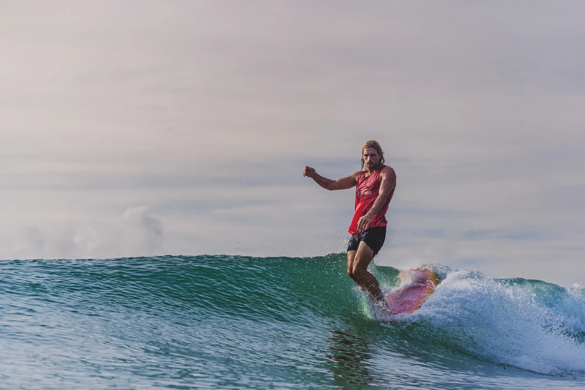 A person with long hair, wearing a red tank top and black shorts, is surfing on a small wave under a cloudy sky. They appear balanced and focused while riding the surfboard.