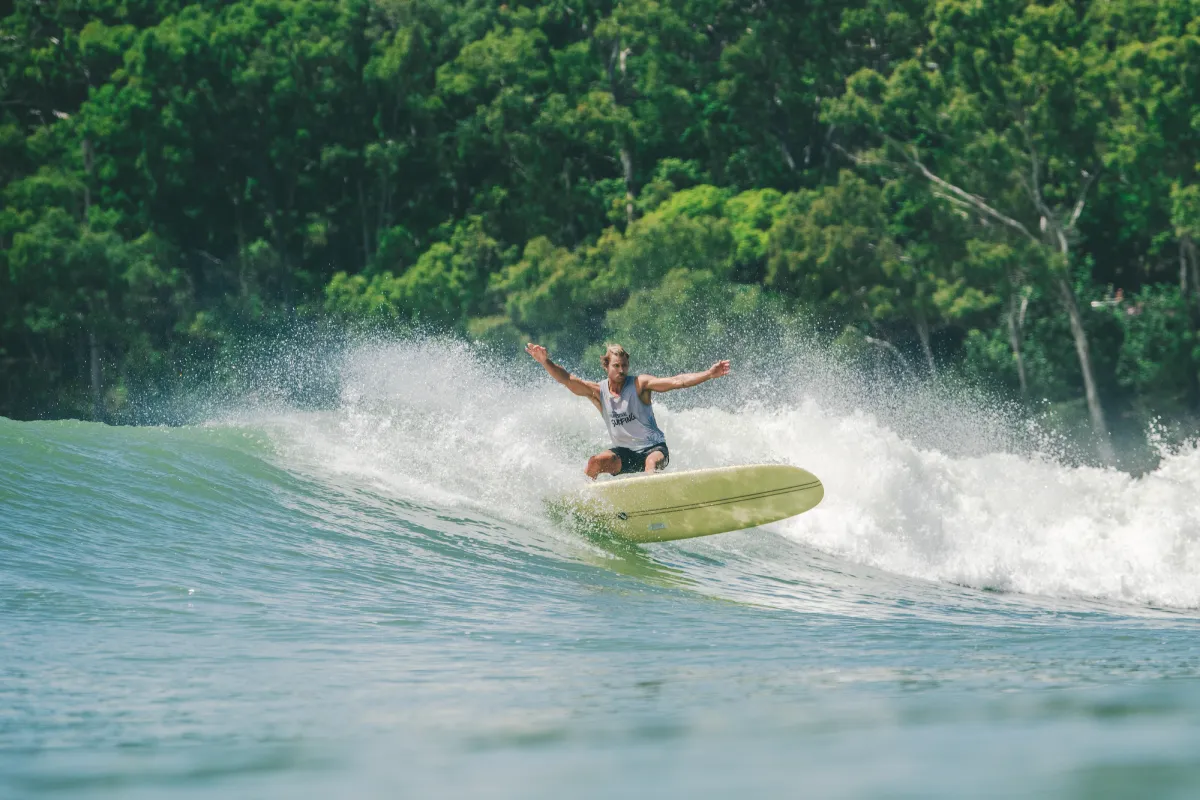 A person surfs on a large wave with arms outstretched, riding a yellow surfboard. Green trees and vegetation are visible in the background under bright sunlight.