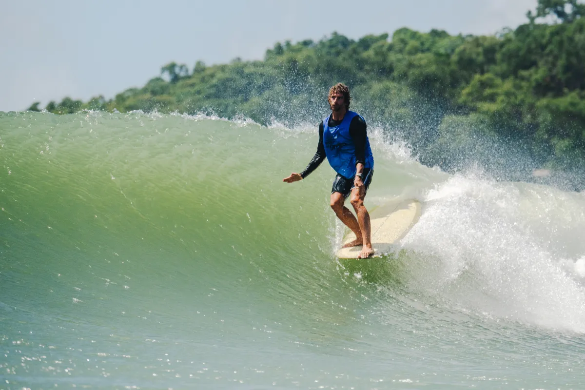 A man in a blue vest and black sleeves surfs on a large green wave near a lush, tree-covered shoreline under a bright sky.