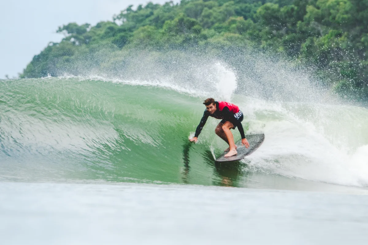 A surfer in a black and red shirt rides inside the barrel of a green wave, crouched low on the surfboard, with lush green trees visible in the background.