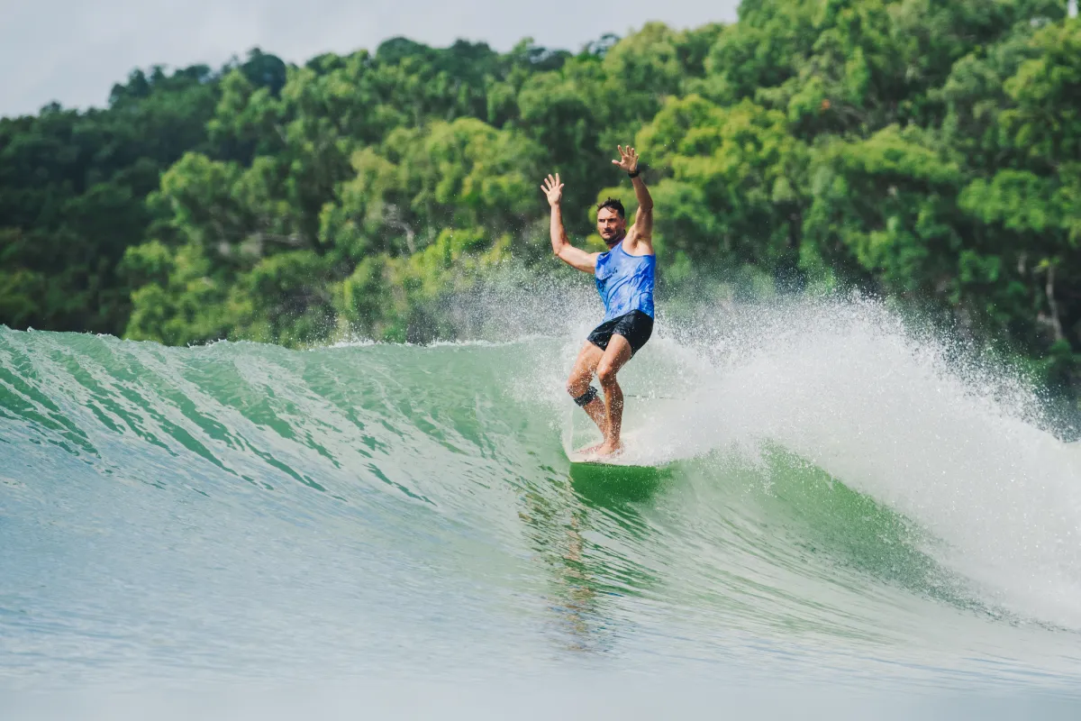 A person in a blue tank top and black shorts is surfing on a large wave with arms raised, surrounded by lush green trees in the background.