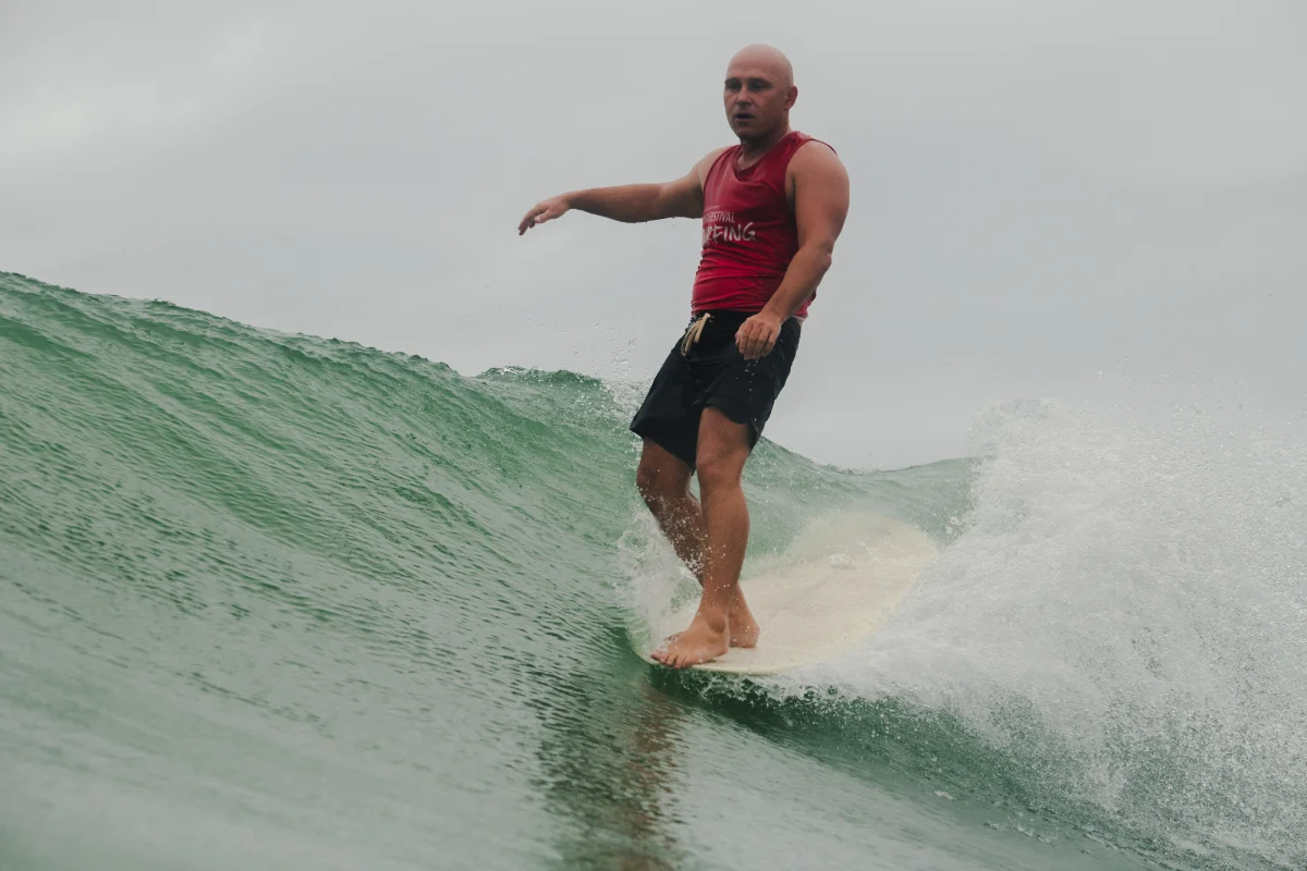 A man wearing a red sleeveless shirt and black shorts is surfing on a green wave, balancing barefoot on a surfboard under an overcast sky.