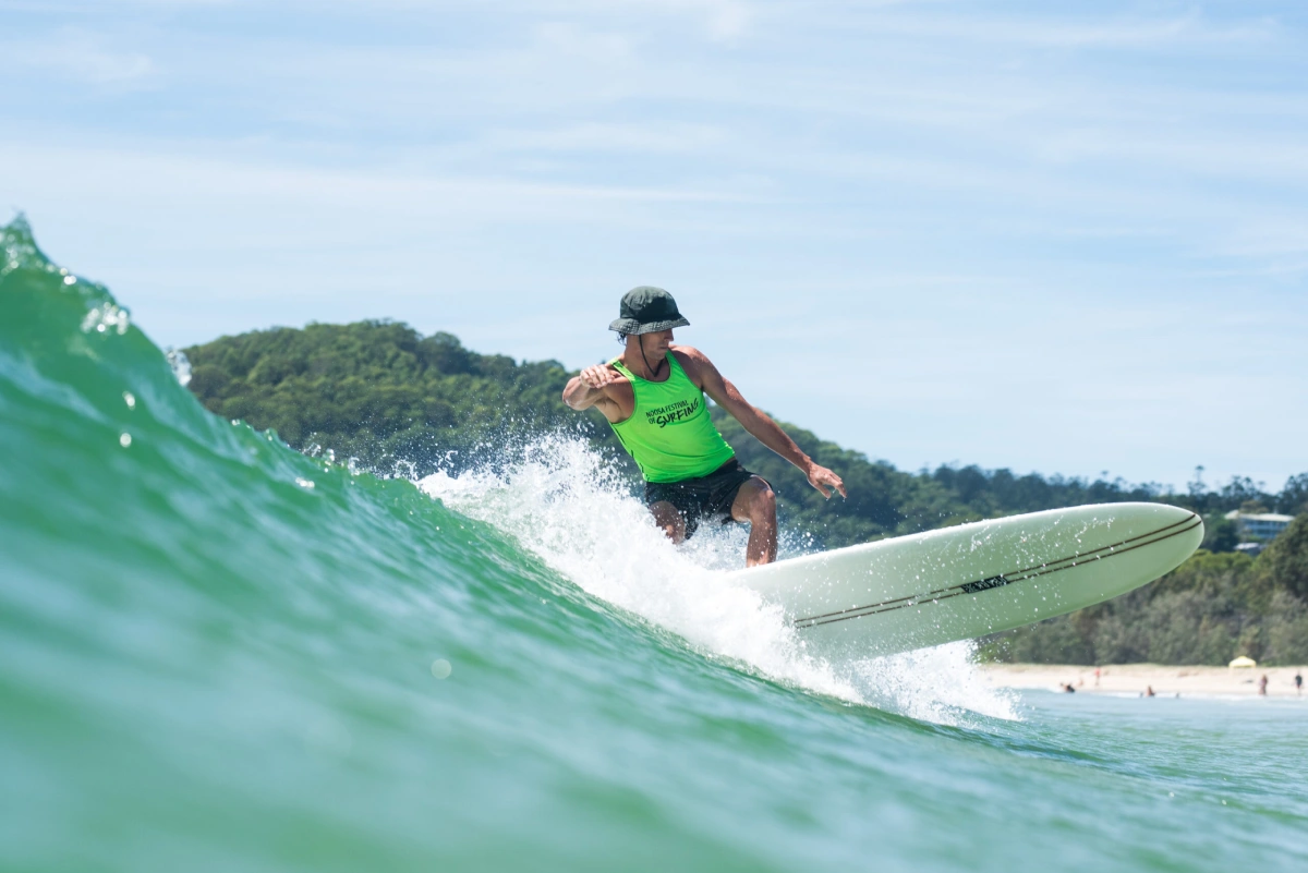 A surfer wearing a green tank top and black hat rides a wave on a white surfboard near a beach, with green hills and trees in the background.