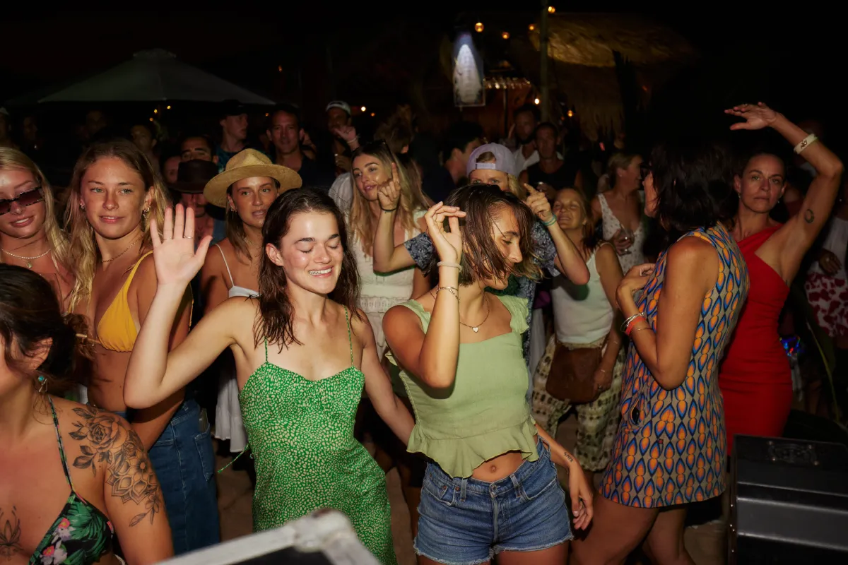 A large group of young adults dance and enjoy themselves at a lively outdoor party at night, some smiling and raising their hands, while others mingle in the background under dim lights.