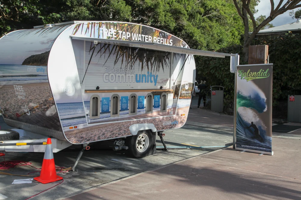 A trailer with a beach scene design offers free tap water refills from five dispensers. A banner with a surfing image and the word "Splendid" stands nearby. The setup is outdoors near trees and pavement.