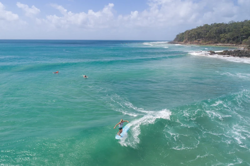 A surfer rides a turquoise wave near a rocky shoreline with trees under a partly cloudy sky. Two other people are floating in the water further out.