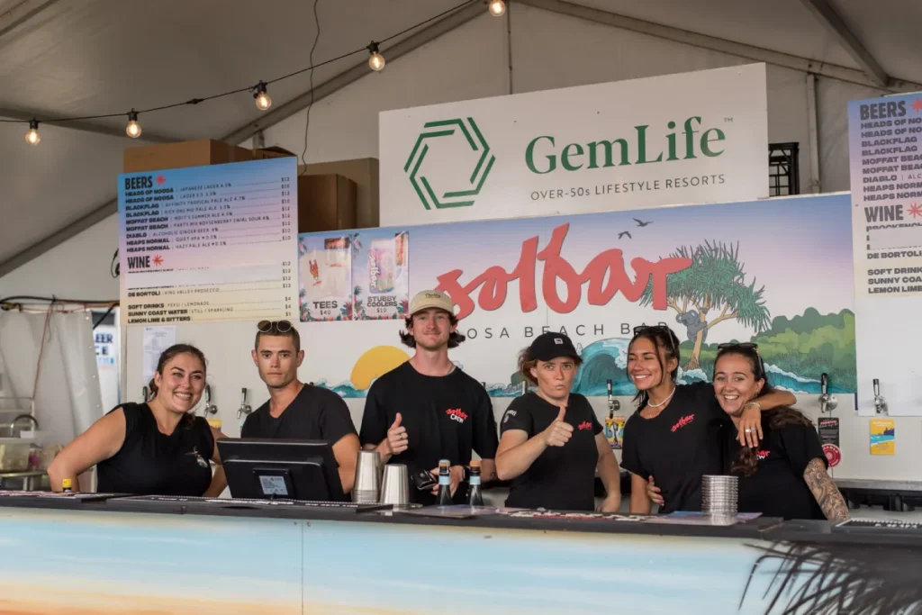 Six smiling staff members stand behind a bar counter under a tent, posing for a group photo. A colorful Solbar sign and GemLife banner are displayed behind them, along with menus and decorative lights.