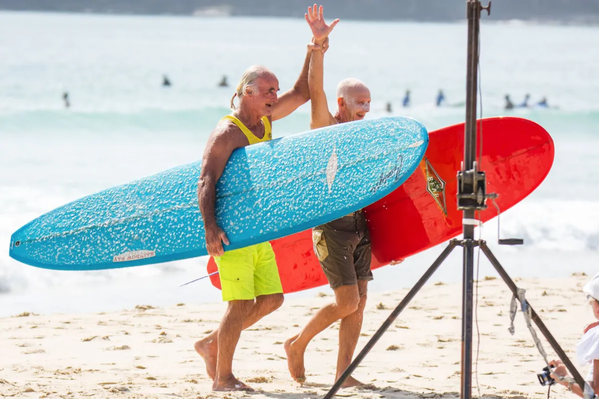 Two older men carrying surfboards walk on a sandy beach; one waves and smiles while the other holds a red board. Both are in swimwear, with the ocean and surfers in the background. A camera tripod is visible in the foreground.