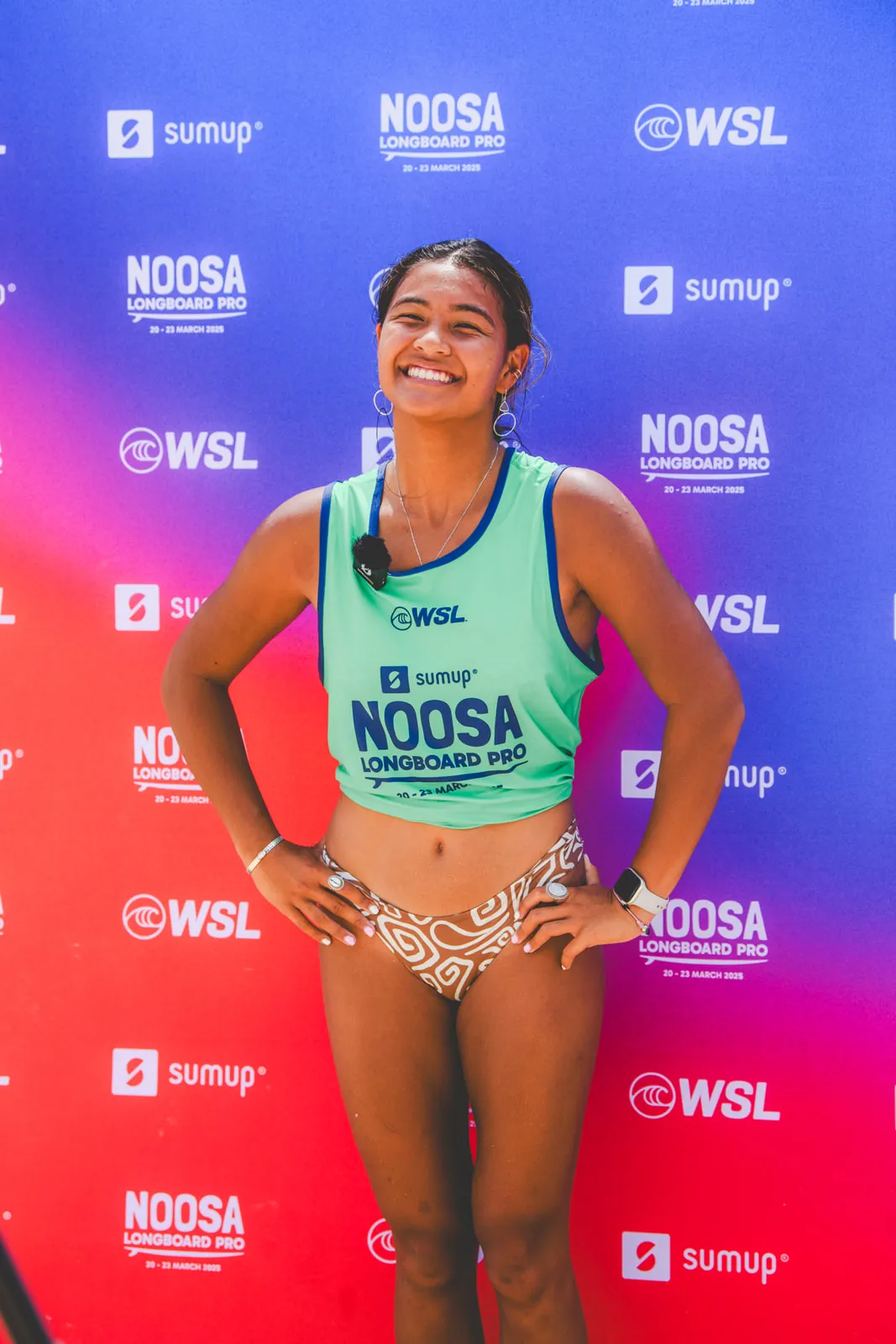 A smiling woman wearing a green "Noosa Longboard Pro" vest and patterned bikini bottoms stands in front of a red and blue event backdrop with WSL and Noosa Longboard Pro logos.