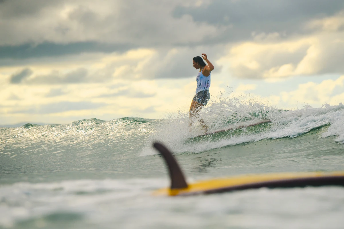 A surfer rides a wave under a cloudy sky, balancing on their board. In the foreground, part of another surfboard is visible in the water. The scene captures dynamic movement and ocean spray.