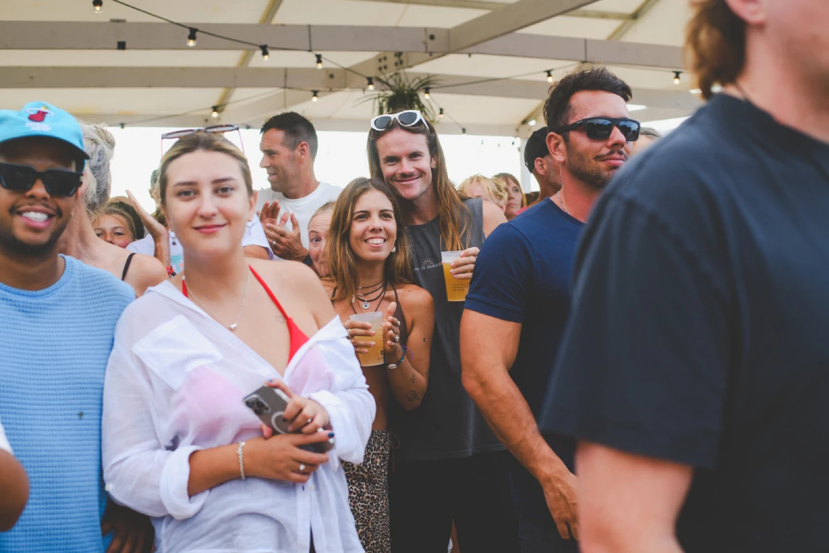 A group of people at an outdoor event smile and enjoy drinks under a tent. The atmosphere is lively, with some people wearing sunglasses and casual summer clothes.