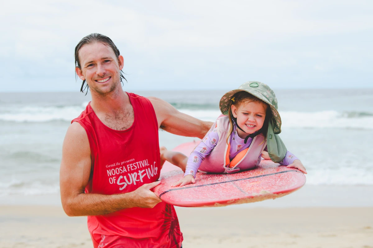 A man in a red "Noosa Festival of Surfing" tank top holds a surfboard with a smiling young child in a hat and wetsuit lying on it at the beach, with the ocean in the background.