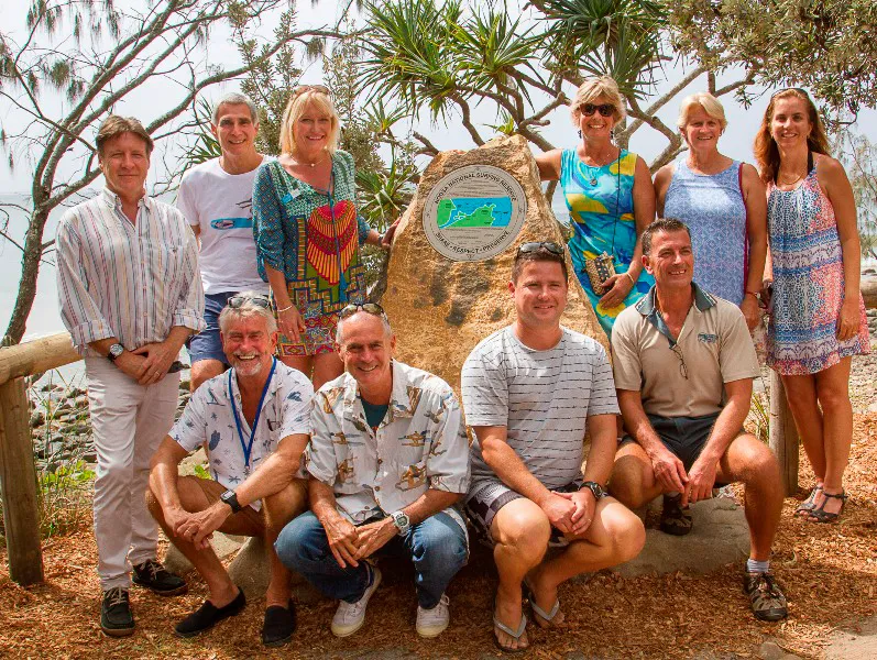 A group of ten adults, five seated and five standing, pose and smile outdoors around a large commemorative stone plaque with greenery and trees in the background.