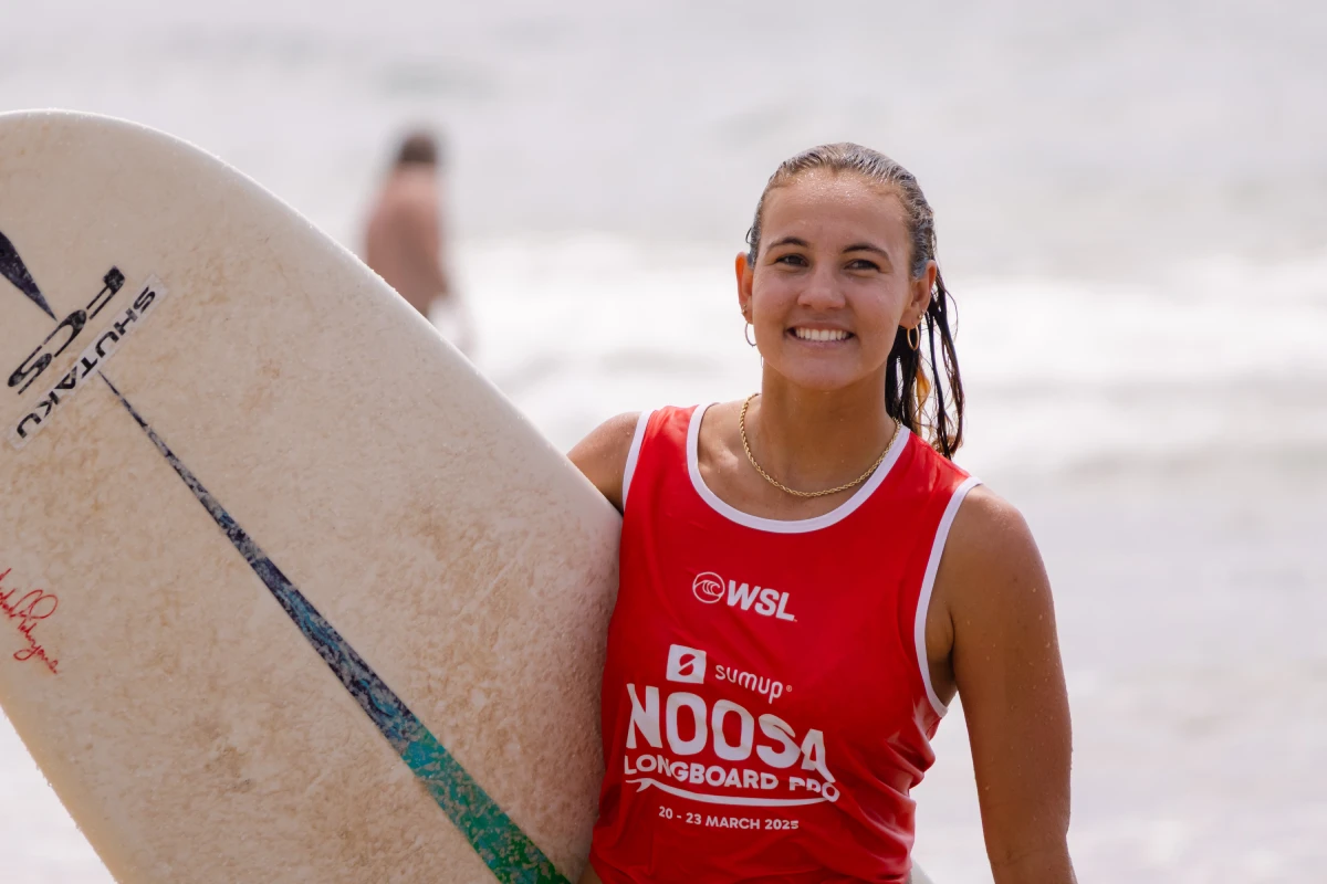 A smiling surfer in a red "Noosa Longboard Pro" tank top stands on the beach holding a surfboard, with the ocean and a blurred figure in the background.
