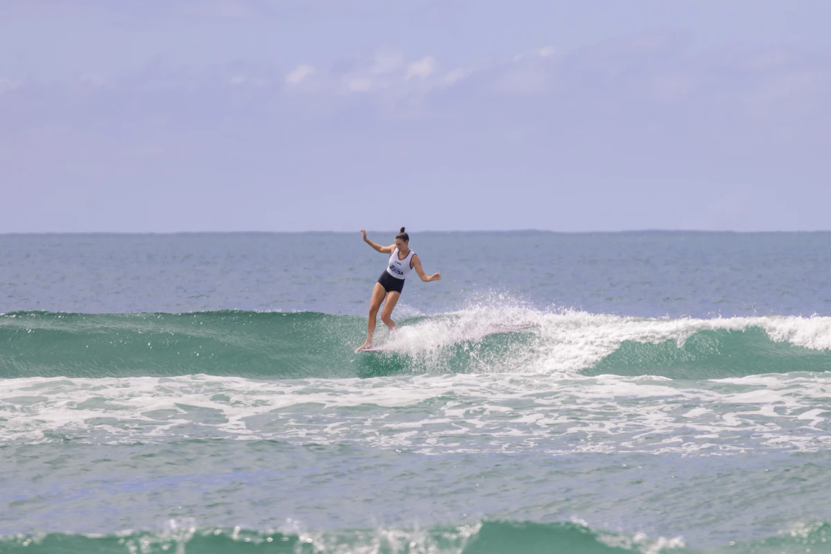 A person in a white shirt and dark shorts is balancing while surfing on a small ocean wave under a partly cloudy sky.