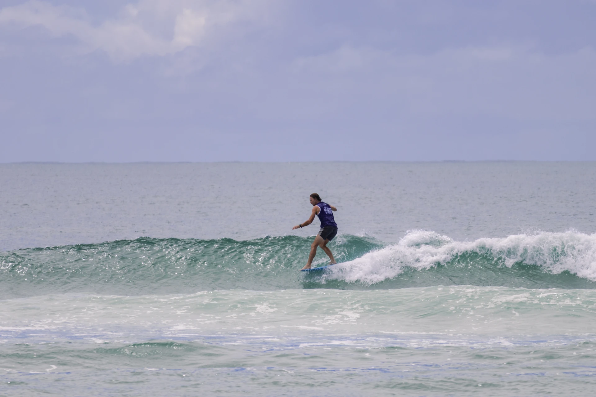 A person wearing a blue shirt and shorts is surfing on a small wave in the ocean under a partly cloudy sky. The water is turquoise and the scene appears calm and relaxed.