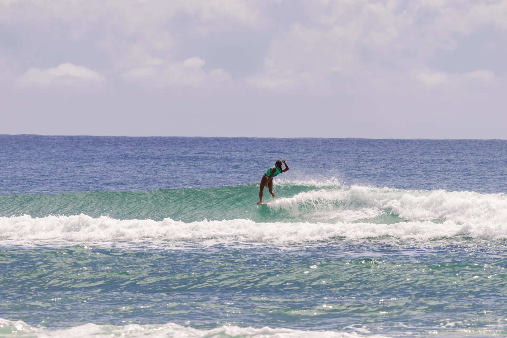 A surfer in a green shirt rides a small wave on the ocean, balancing on their surfboard with one arm raised, under a bright sky with scattered clouds.