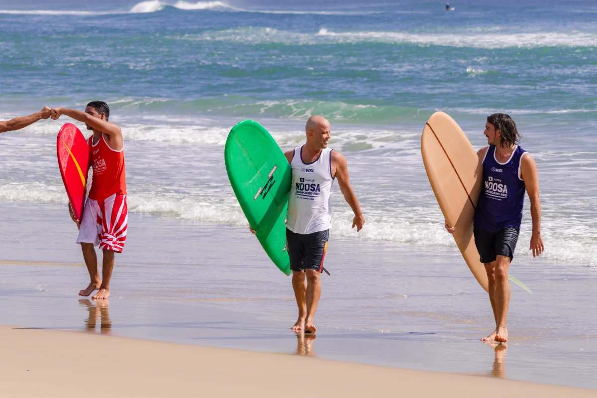 Three men carrying surfboards walk along the beach. Two are wearing white tanks, one in a dark tank, and one gives a fist bump to someone off-frame. Waves roll in behind them under a clear sky.
