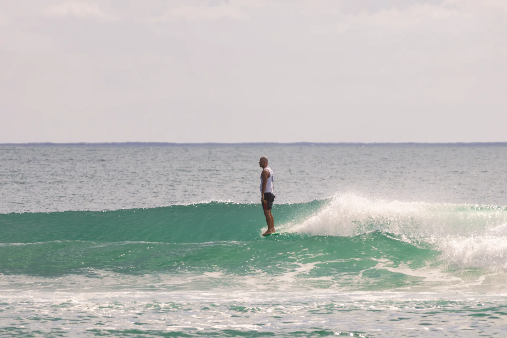 A person stands upright on a surfboard, riding a gentle turquoise wave in the ocean under a mostly cloudy sky.