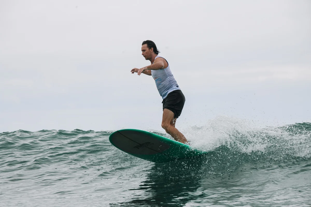 A person wearing a light tank top and black shorts rides a green surfboard on a small ocean wave under a cloudy sky.