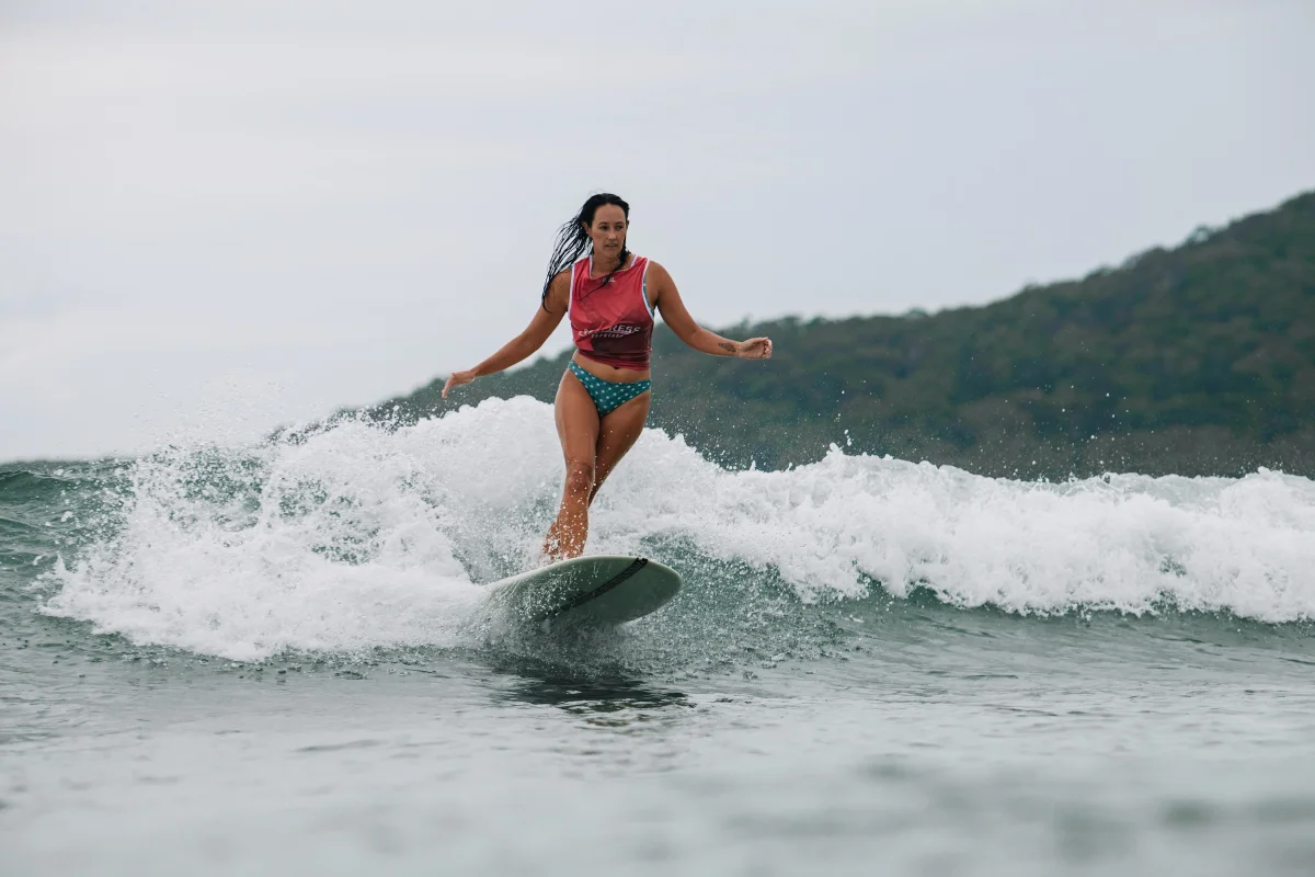 A person in a pink shirt and blue shorts surfs on a wave with green hills and a cloudy sky in the background.
