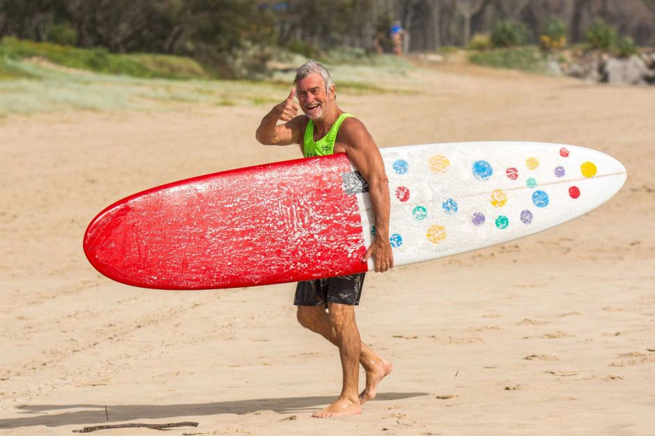 An older man in a green tank top smiles while carrying a surfboard with a red tip and colorful dots, walking barefoot on a sandy beach with greenery in the background.