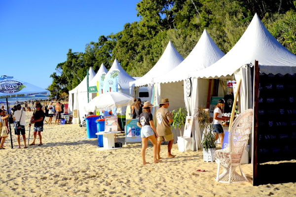 A row of white tents set up on a sunny beach, with people walking between the stalls and others gathered under umbrellas. Green trees line the background.