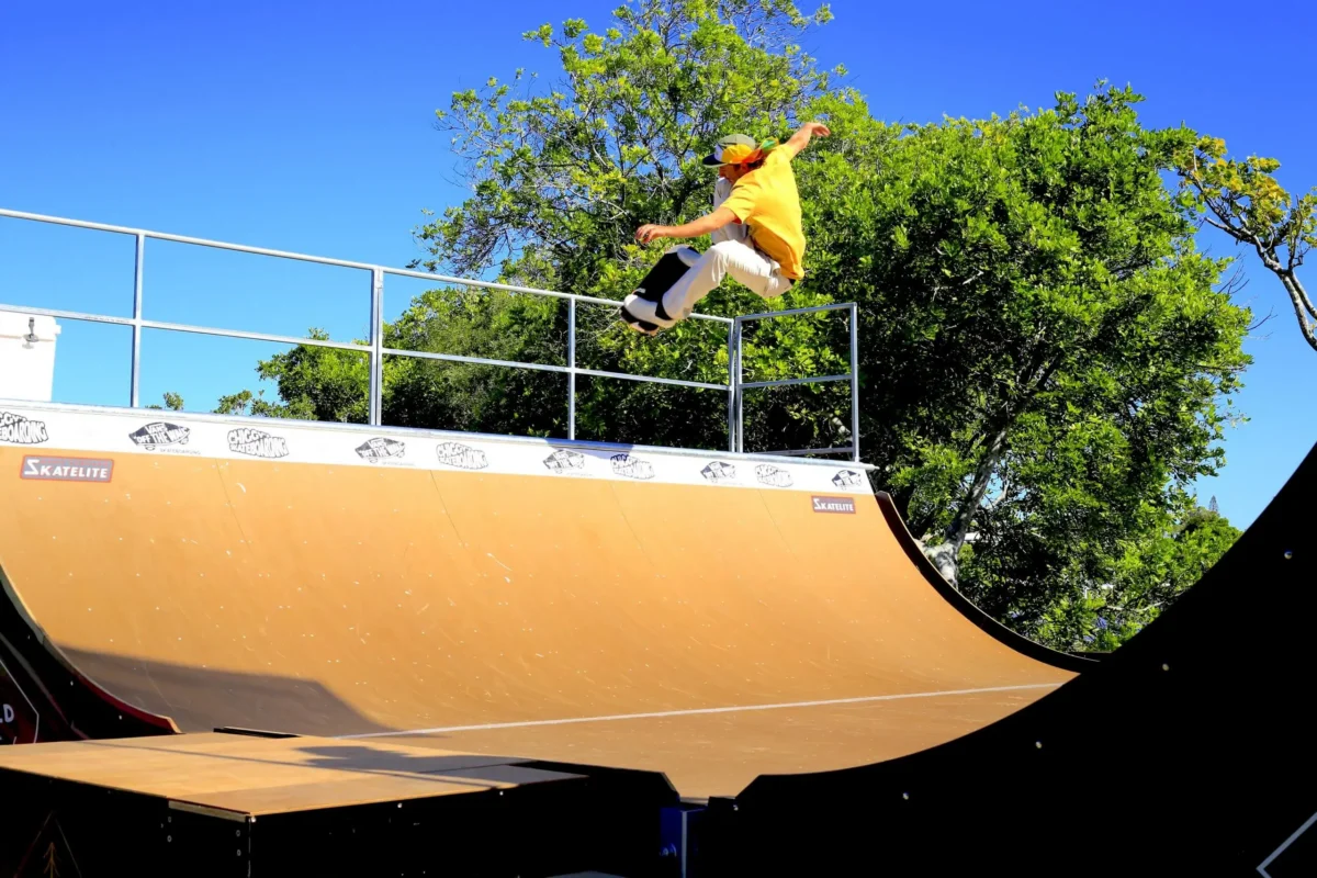 A skateboarder in a yellow shirt and cap performs a high jump trick on a half-pipe ramp outdoors, with green trees and a clear blue sky in the background.