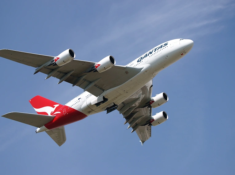 A Qantas airplane with four engines and a red tail featuring a white kangaroo is flying in a clear blue sky, viewed from below.